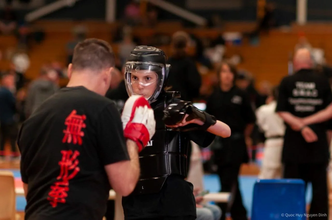 Young martial arts student in black protective gear spars with a coach at a Martial Arts tournament; red Chinese characters on the coach's shirt.