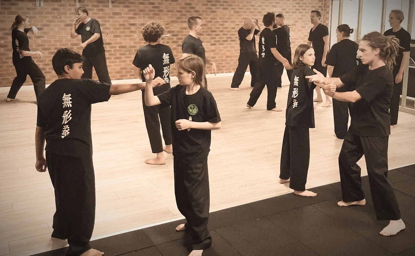 Martial arts class with children and adults practicing partner drills in black uniforms in a studio with a brick wall and Japanese characters on shirts.