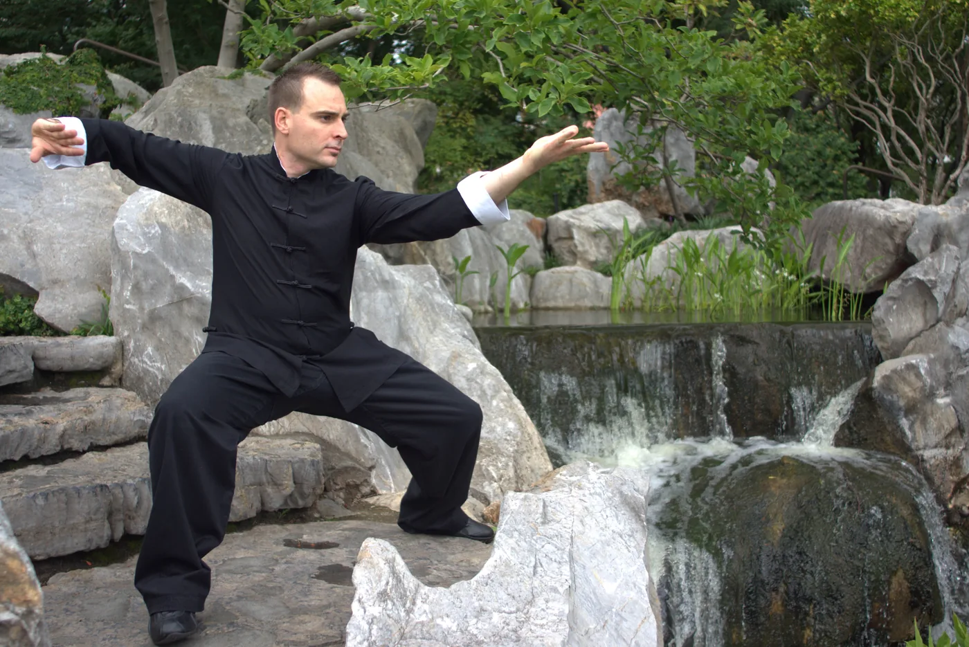 Man in black tai chi outfit holds a focused stance on rocks beside a small waterfall in a lush garden.