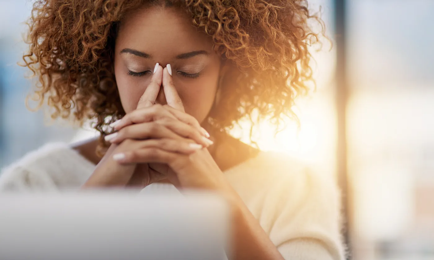Curly-haired woman with eyes closed, pressing fingers to her nose, a clear sign of fading concentration, by a sunlit window.