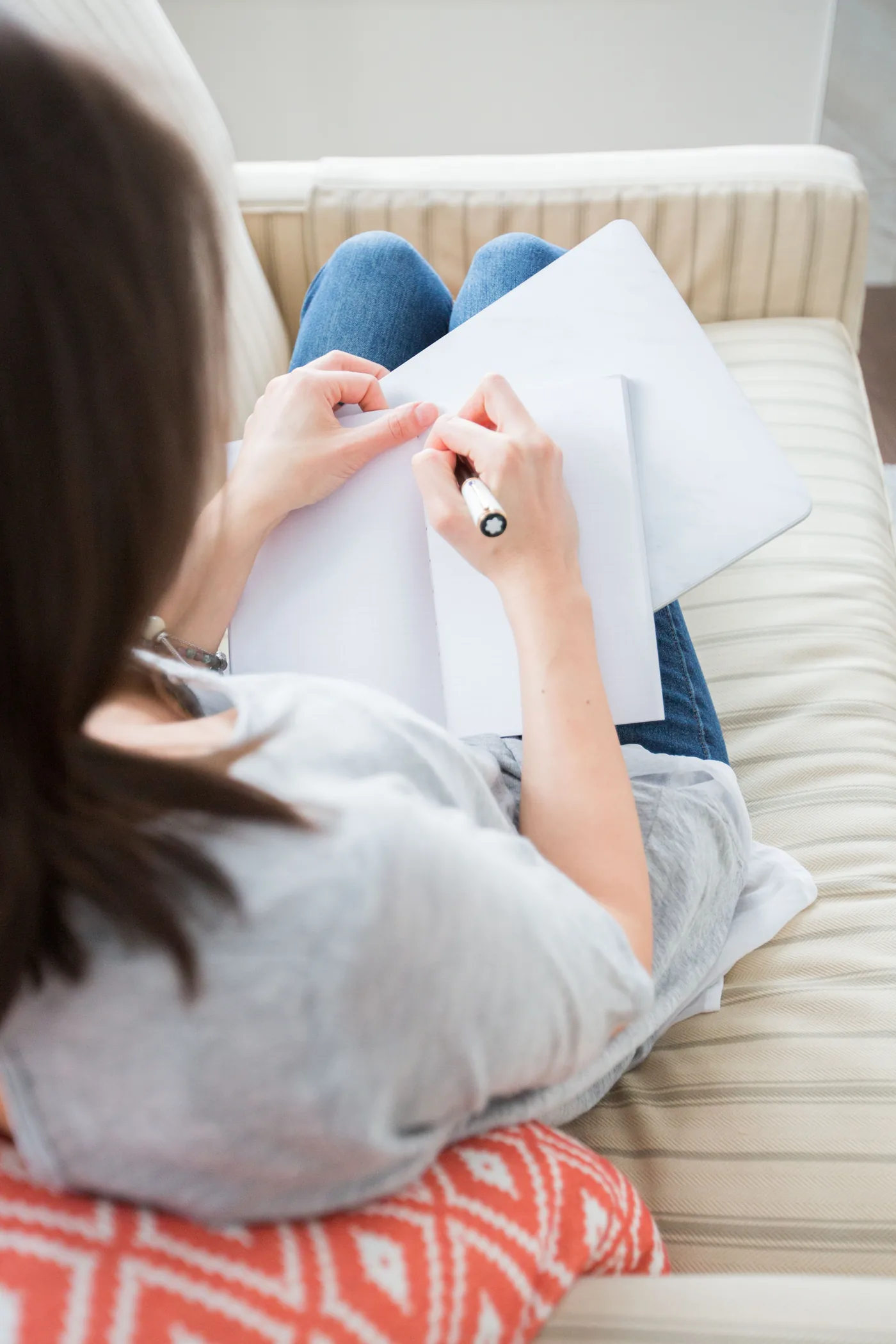 Person sitting on a striped couch writes in a blank notebook, legs crossed, in a calm, bright living room.