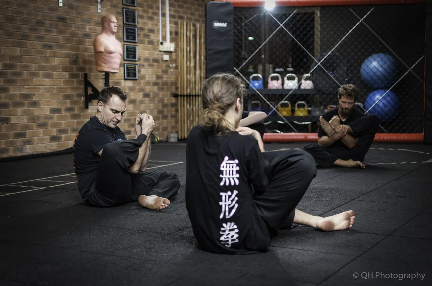Martial arts class seated on gym mats, three people stretching in black uniforms with Chinese characters, brick wall and training gear behind.