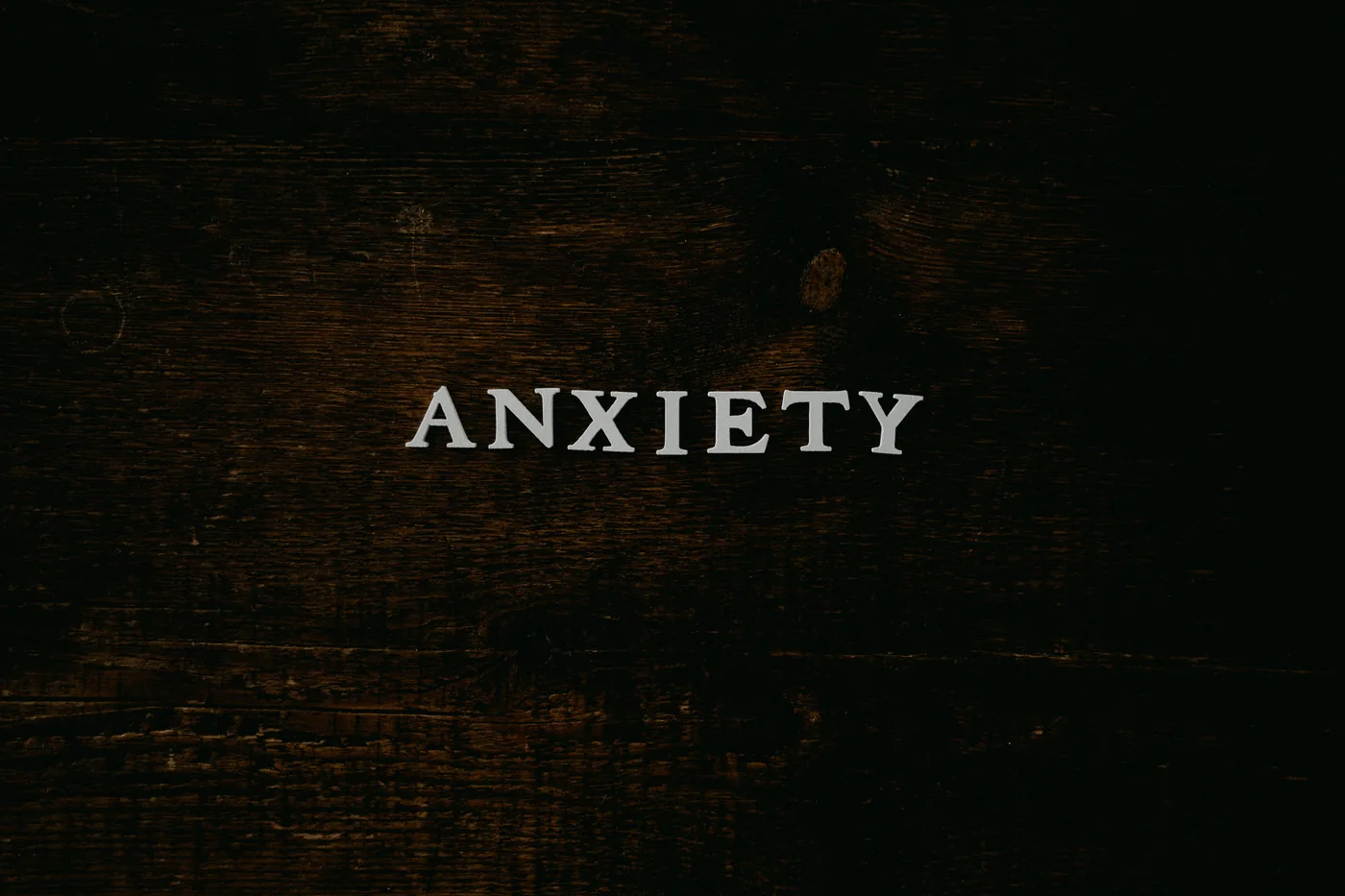 Word ANXIETY in white serif letters on a dark, textured wood background, creating a tense, moody feel.