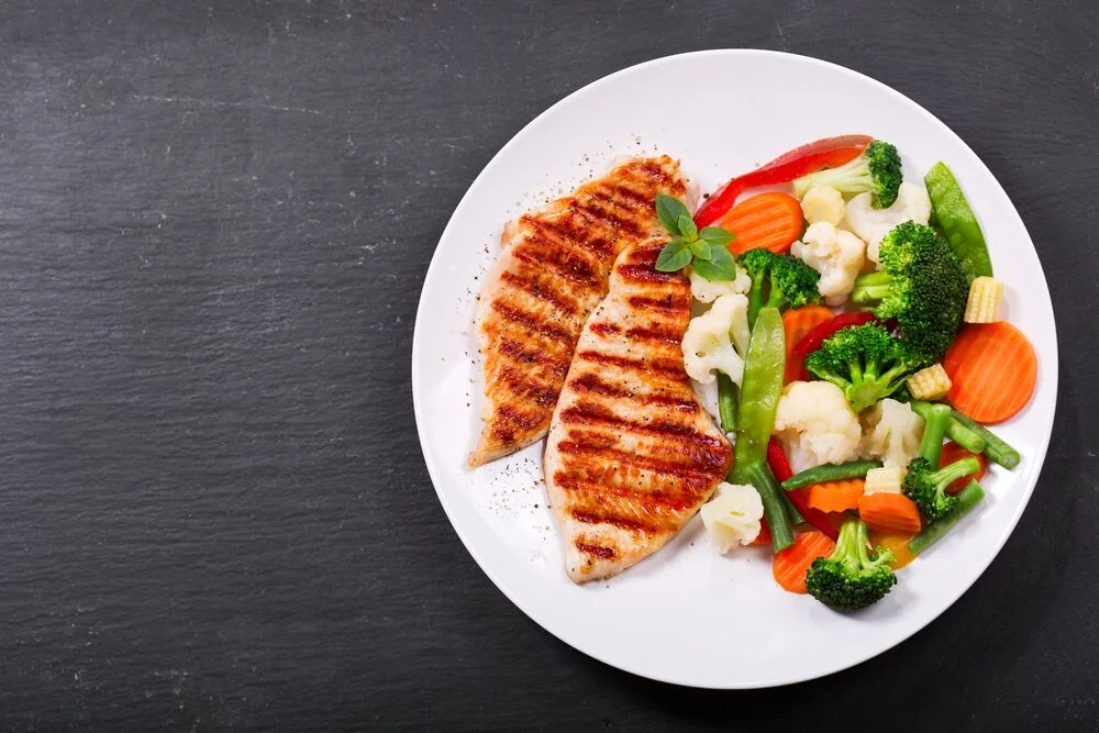 Grilled chicken breasts with mixed vegetables on a white plate against a dark tabletop, a colorful healthy meal.