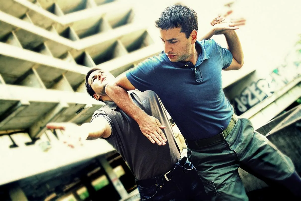 Two men practice self-defense under a concrete building, one blocking the other in an intense pose.