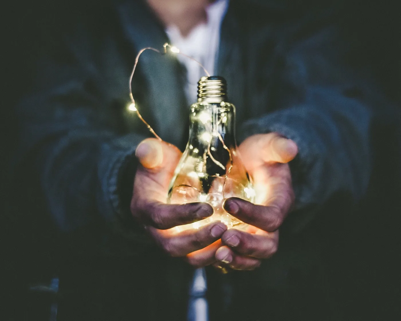 Hands cradle a glowing light bulb wrapped with tiny string lights against a dark, blurred background, creating a warm mood.