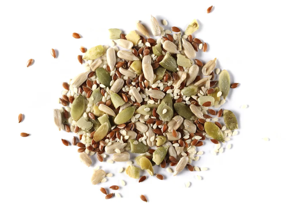 Mixed seeds and grains piled on a white background, with tan, green, brown, and white pieces scattered around them
