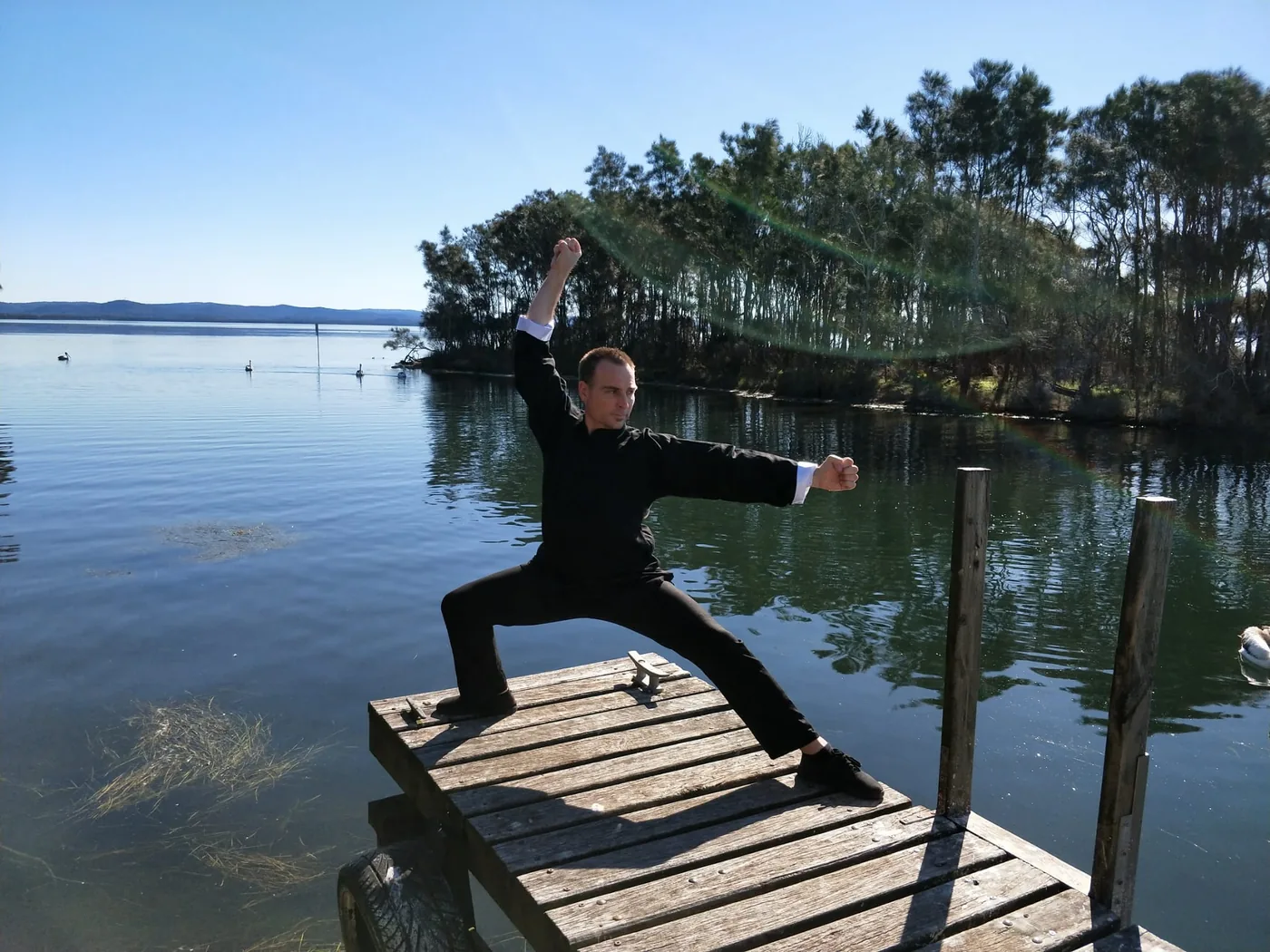 Man in black martial arts pose on a wooden dock by a calm lake, with trees and clear blue sky in the background.