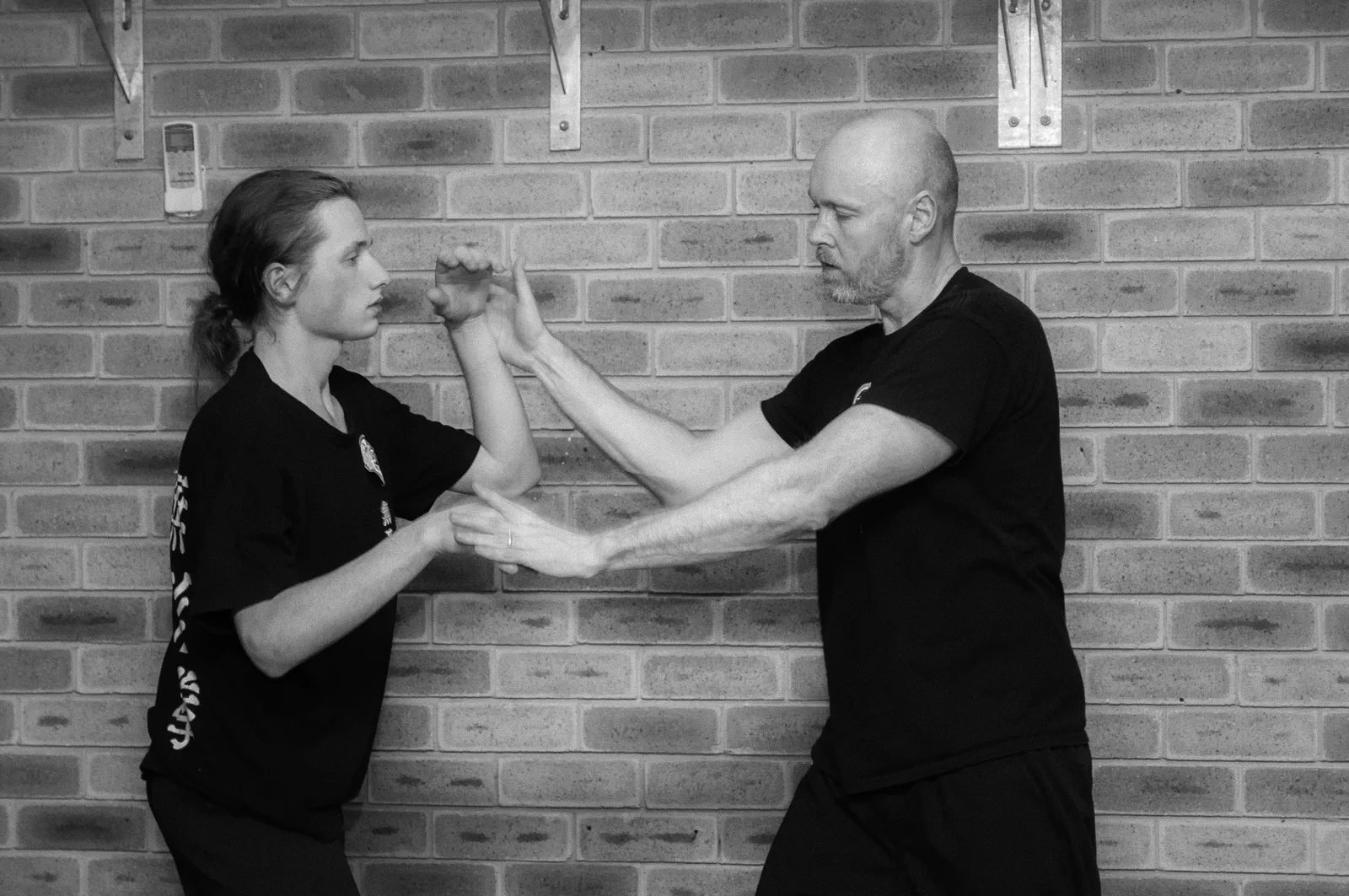 An instructor and a student practise sticky hands against a brick wall, in black and white.
