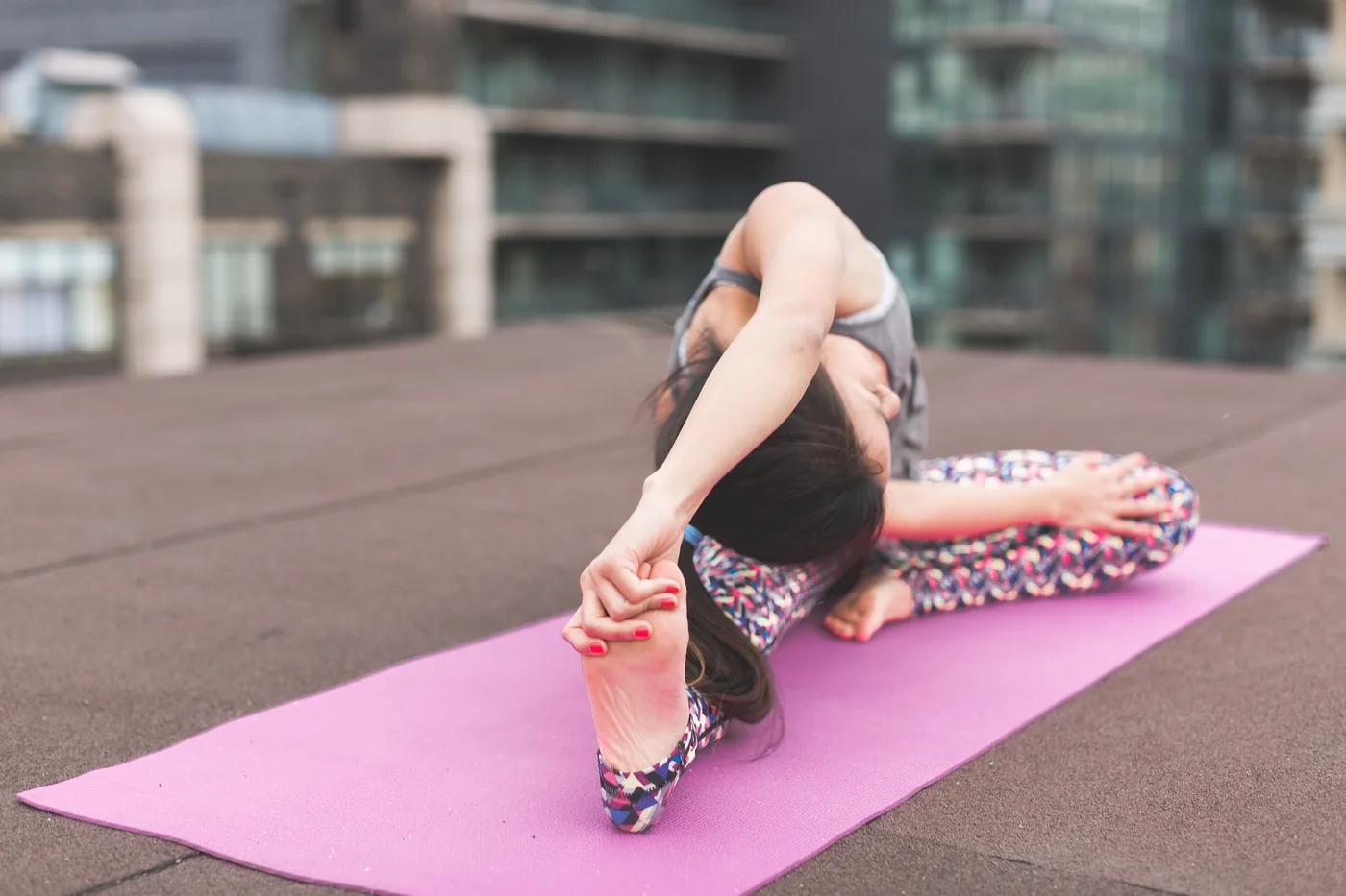 Woman stretches into a yoga pose on a pink mat on a rooftop, with blurred apartment buildings behind her.