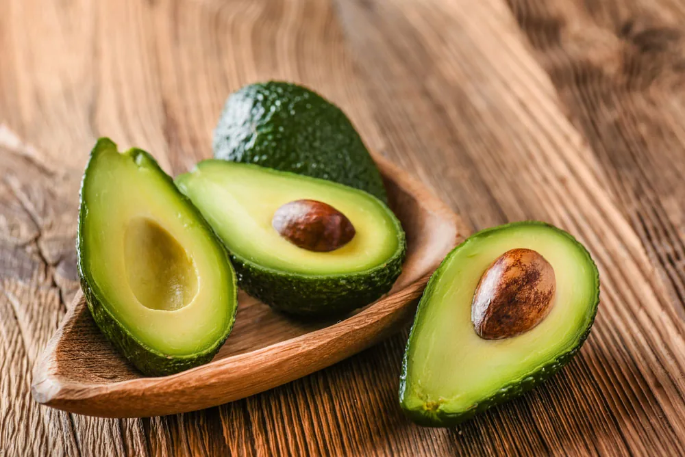 Halved avocados with pits on a wooden spoon and table, showing bright green flesh and rustic kitchen styling.