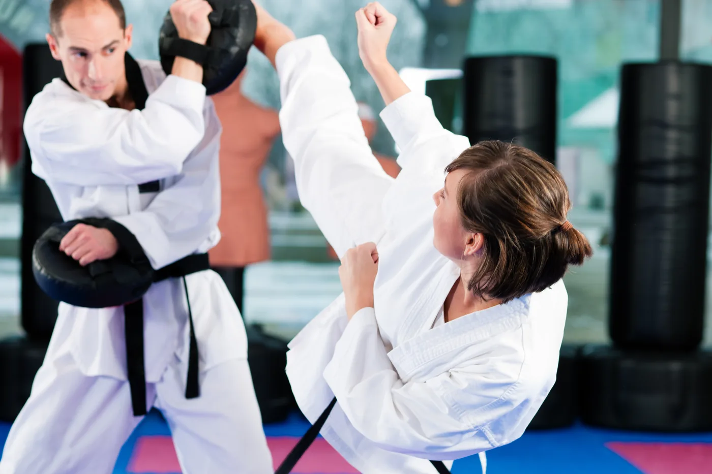 Two karate students in white gis spar on mats, one kicking high while the other blocks with a focus pad in a bright dojo
