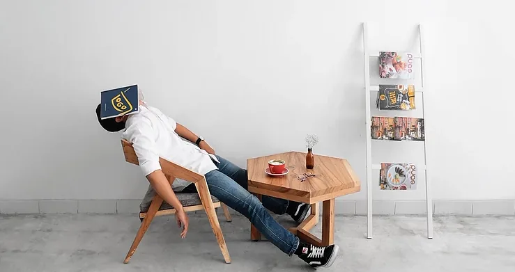 Person asleep in a wooden chair with a book over their face beside a small table with coffee in a sparse white cafe.