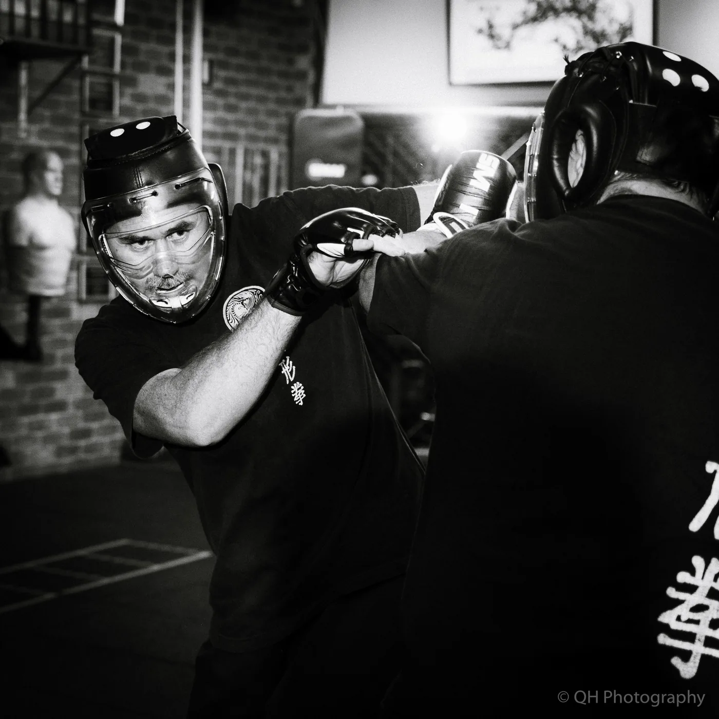 Two helmeted boxers spar in a gym, one blocking a punch; black-and-white scene with intense focus and a QH Photography watermark.