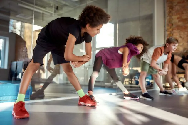 Kids stretch in a bright dance studio, led by a boy in black and red shoes, with others leaning side to side.