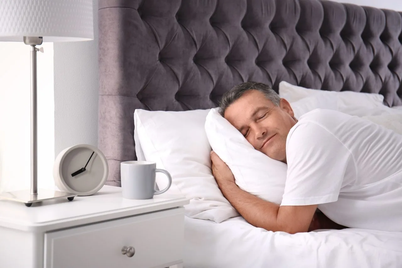 Sleeping man in a white shirt hugs a pillow in a bed with a tufted gray headboard; nightstand holds a lamp, clock, and mug.