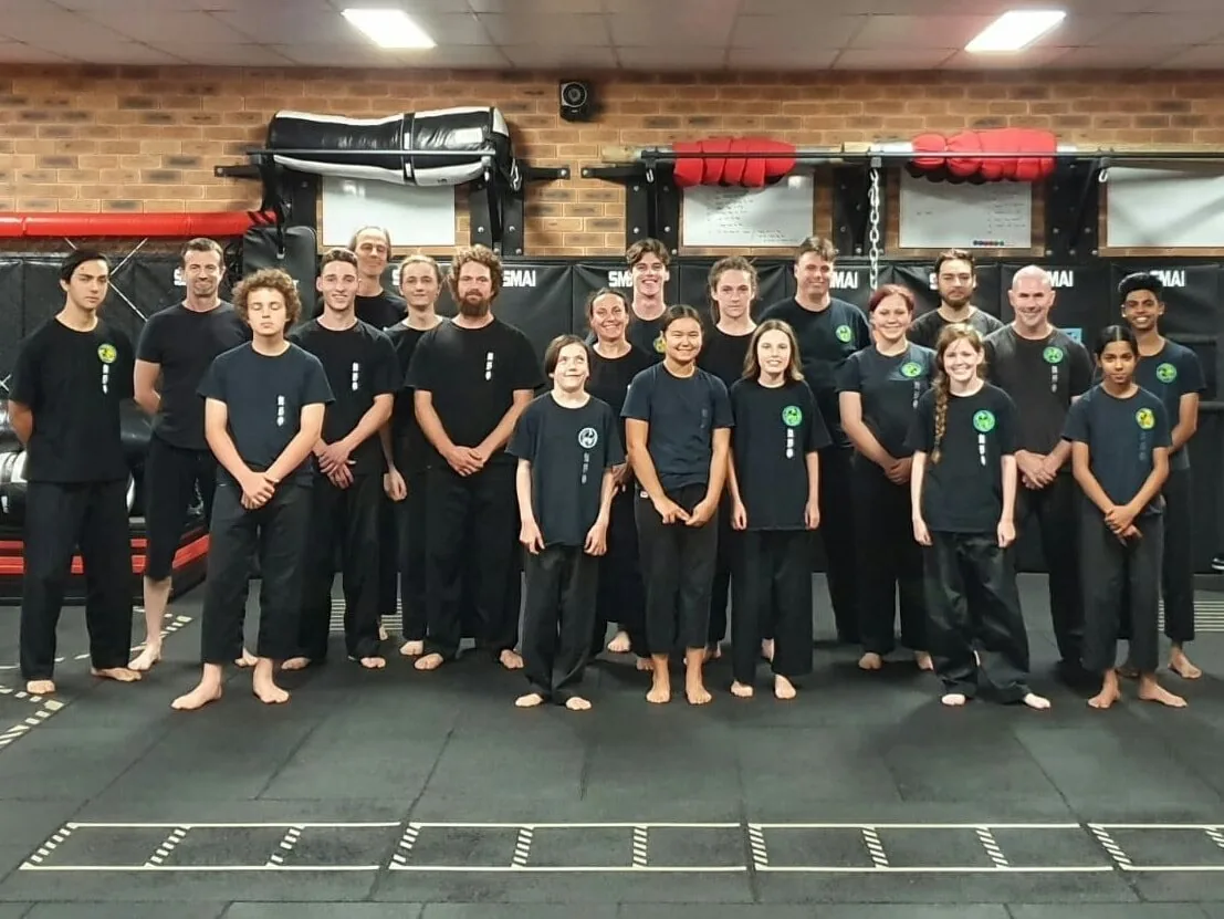 Smiling martial arts students and instructors in black uniforms pose in a dojo with mats, brick wall, and SMMAI banners.