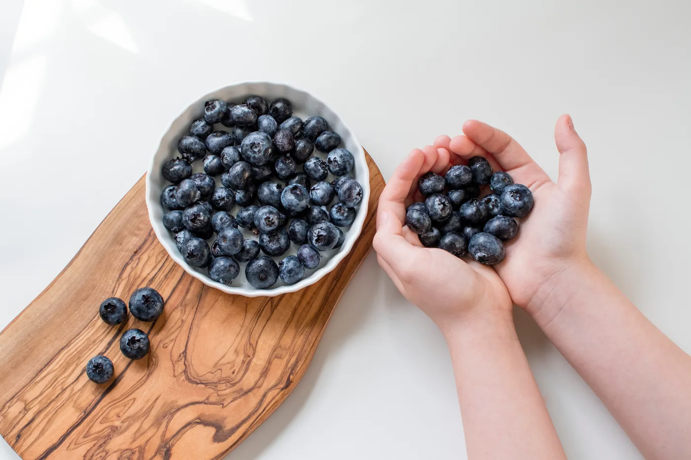 Child’s hands holding blueberries beside a white bowl full of blueberries on a wooden board against a white background.