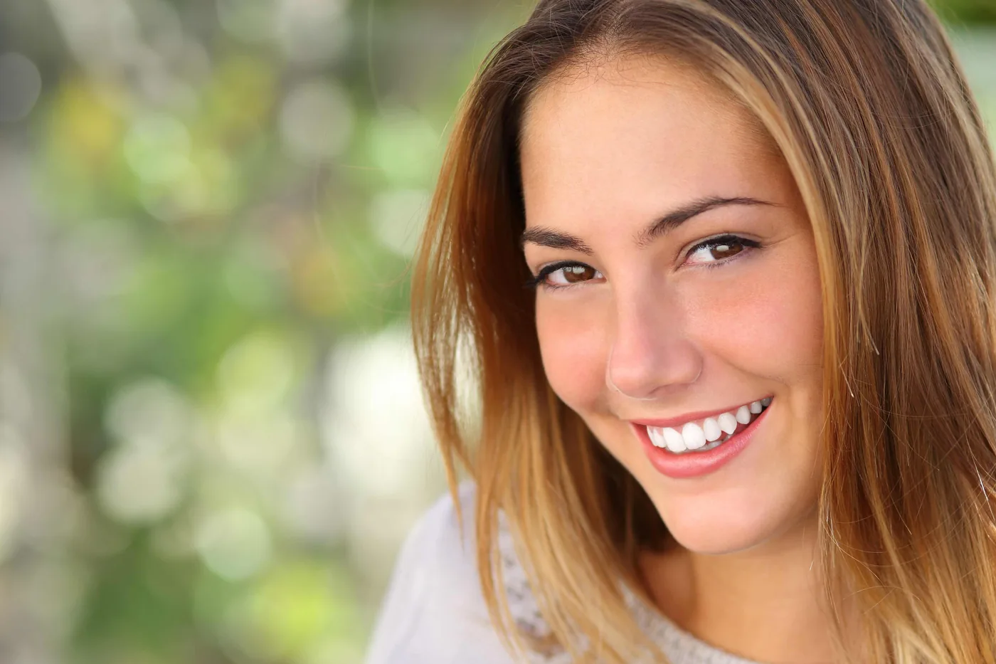 Smiling young woman close-up outdoors with blurred green background, warm light, and long brown-blonde hair.