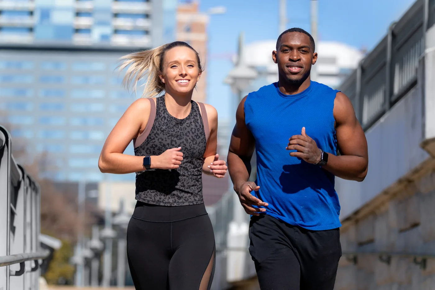 Two smiling runners, a woman in black and a man in blue, jog on a sunny city bridge with buildings blurred behind them.