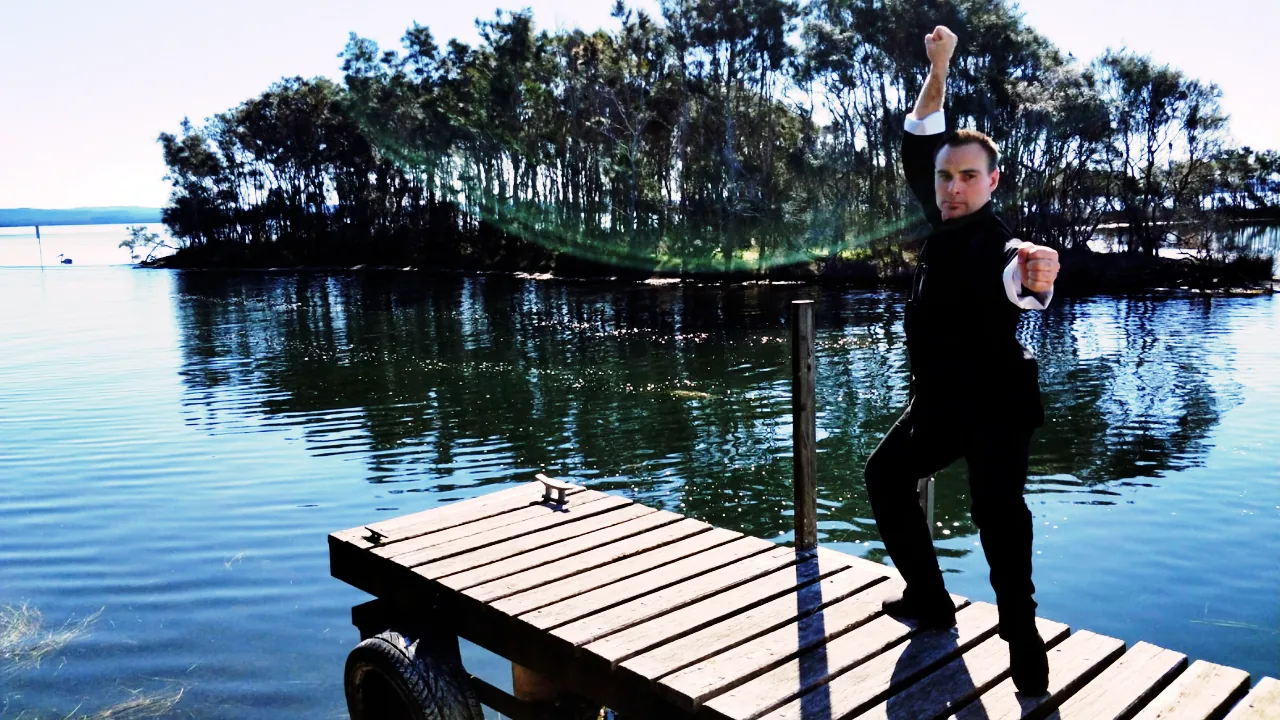 Man in black martial arts pose on a wooden dock by a calm lake, with trees reflected in the water under bright sunlight.