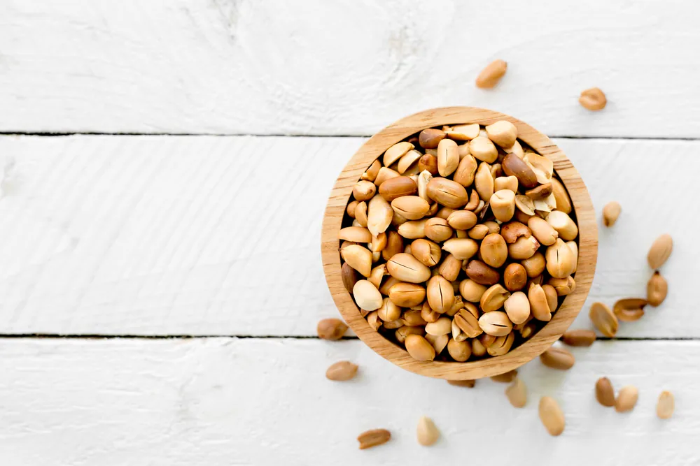 Top view of a wooden bowl filled with peanuts on a white wooden table, with a few scattered around.