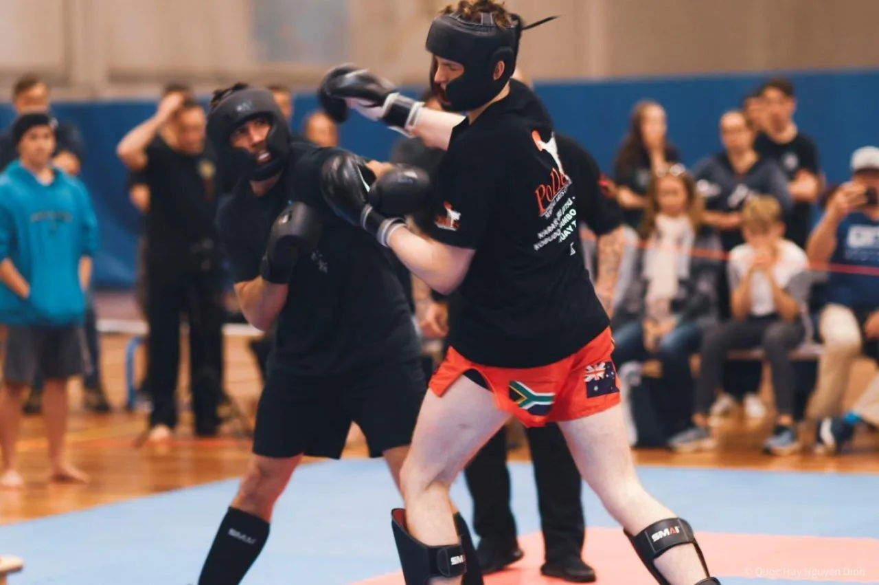 Two helmeted kickboxers spar in an indoor gym while spectators watch; one wears red shorts with flags, the other black.