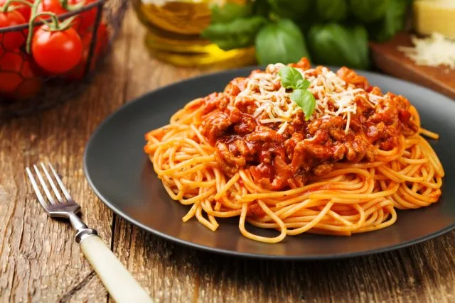 Plate of spaghetti with meat sauce, basil and grated cheese on a dark dish, with a fork and tomatoes on a rustic wooden table.