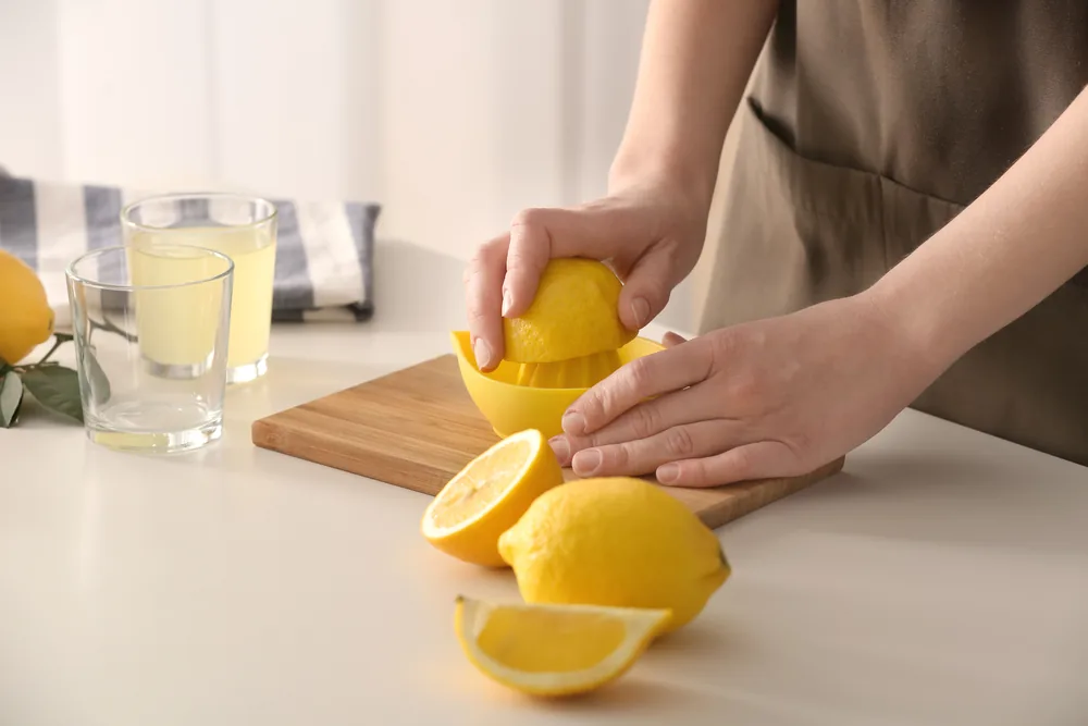 Hands squeezing a lemon in a yellow juicer on a wooden board, with lemon halves and two glasses of lemonade in a bright kitchen