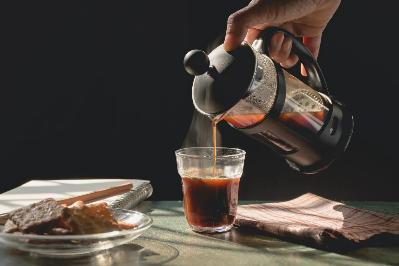 Hand pours hot coffee from a French press into a glass beside pastries on a sunlit table, with a dark background and steam.