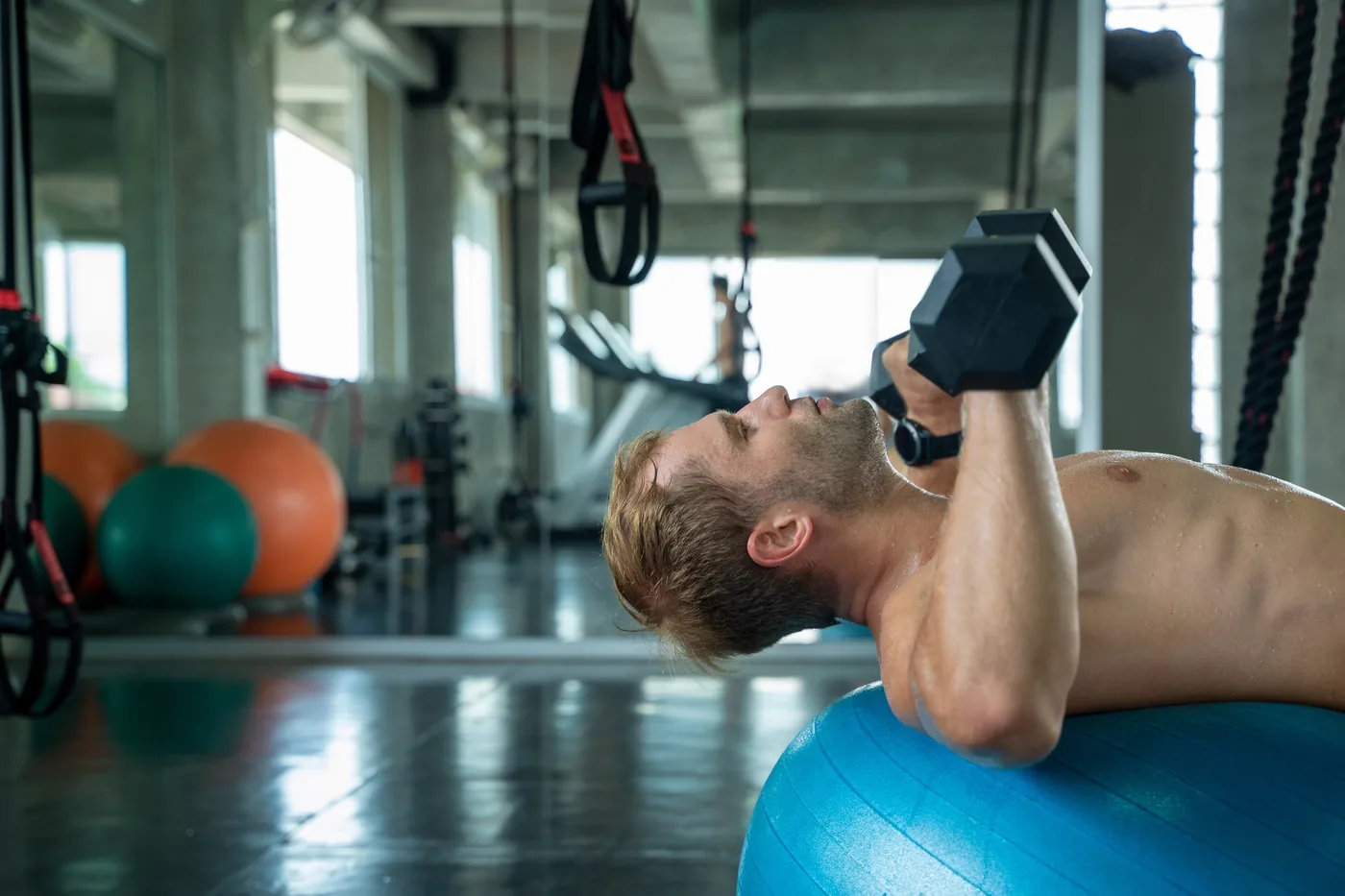 Man lifting dumbbells on a blue stability ball in a gym, with exercise balls and straps in the background.