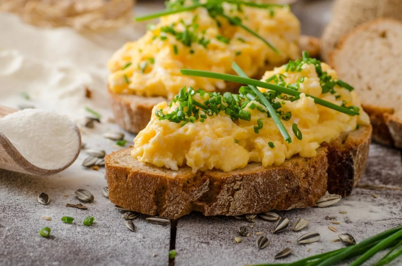 Scrambled eggs with chives on rustic bread slices, scattered sunflower seeds and a wooden spoon on a rustic table.