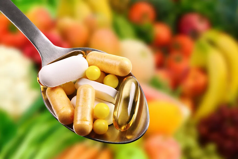 Spoonful of assorted pills and capsules in front of a blurred colorful fruit and vegetable background