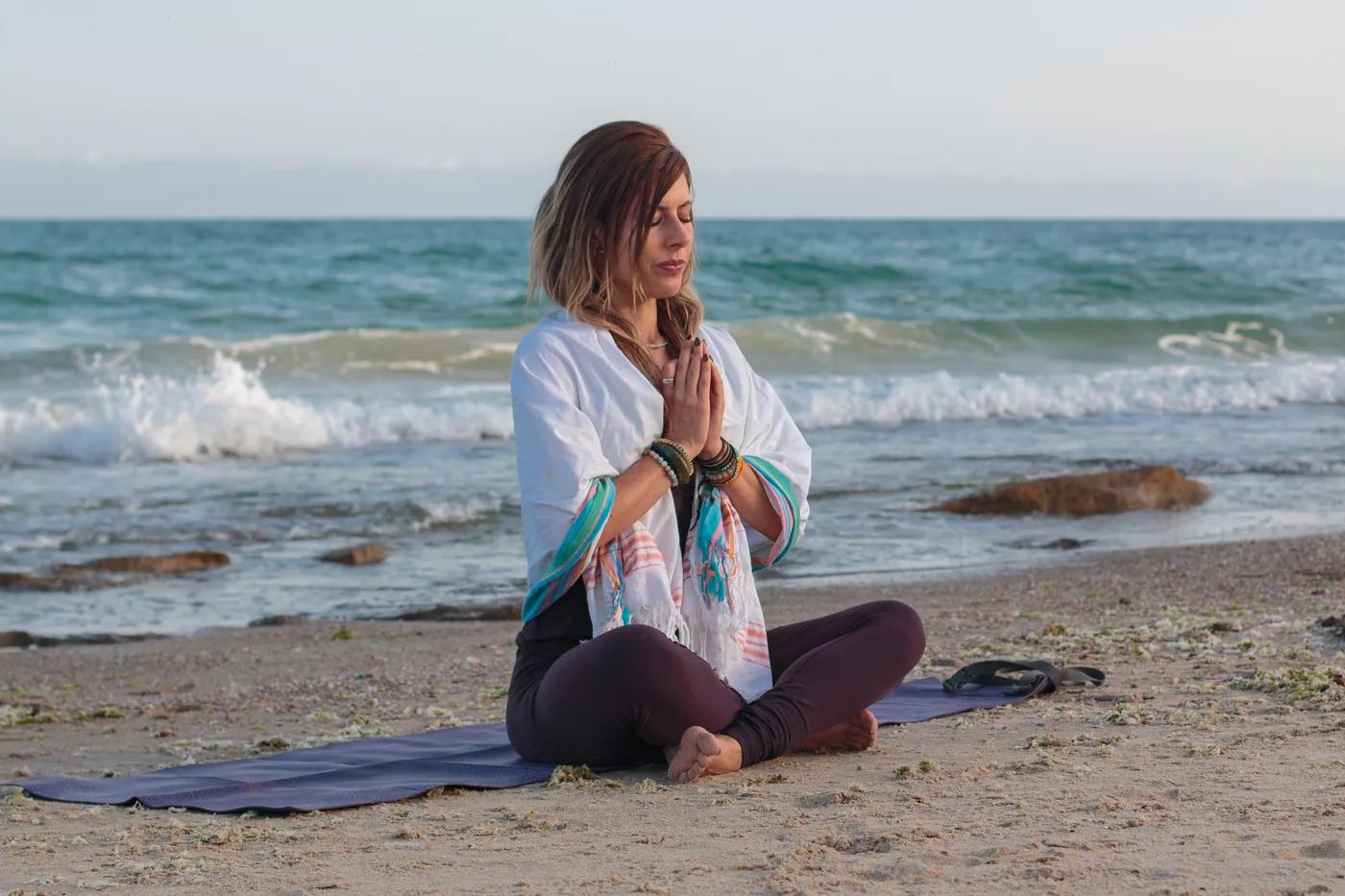 Woman meditating cross-legged on a beach mat, hands in prayer, with waves and rocks behind her under a calm sky.