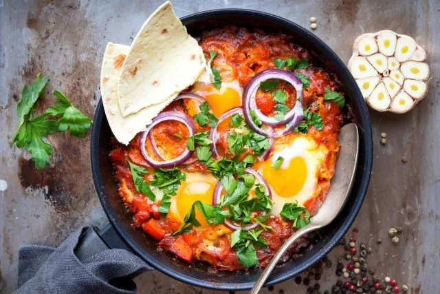 Skillet shakshuka with eggs, red sauce, onion rings, herbs and pita on a rustic table, with garlic and a spoon.