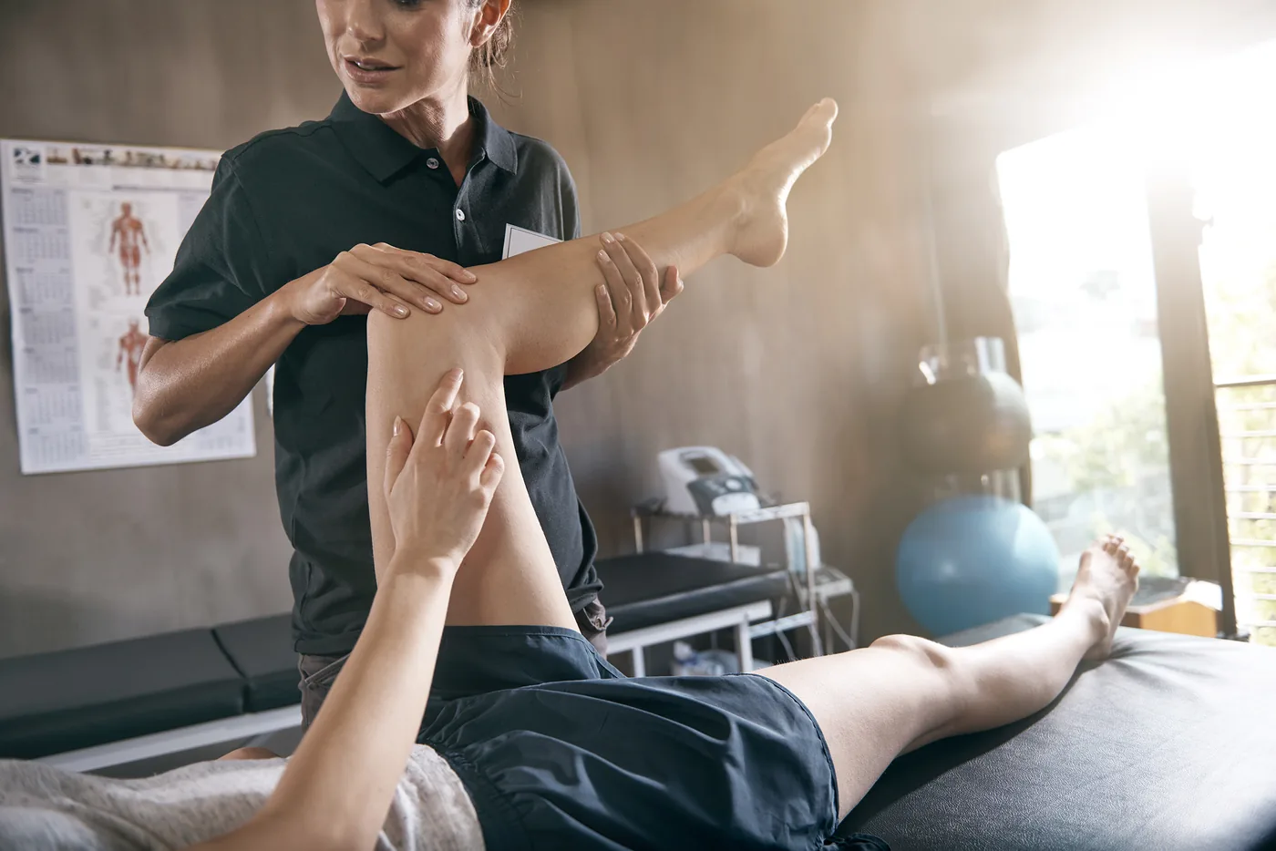 Female therapist stretches a patient’s leg on a clinic table, with anatomy posters and bright window light in the background.