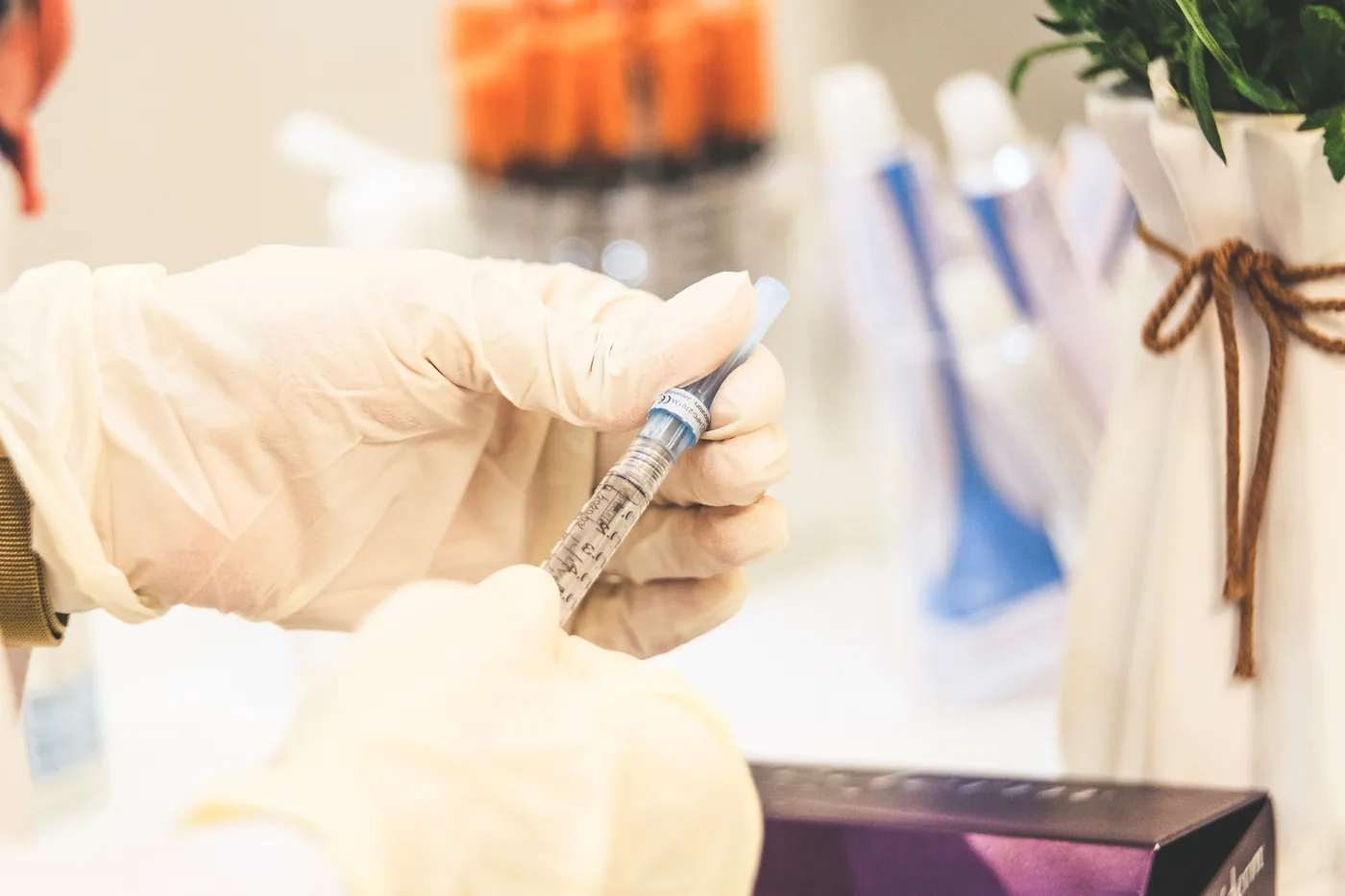 Gloved hands hold a syringe near a blue vial in a bright clinic, with blurred bottles and a potted plant in the background.