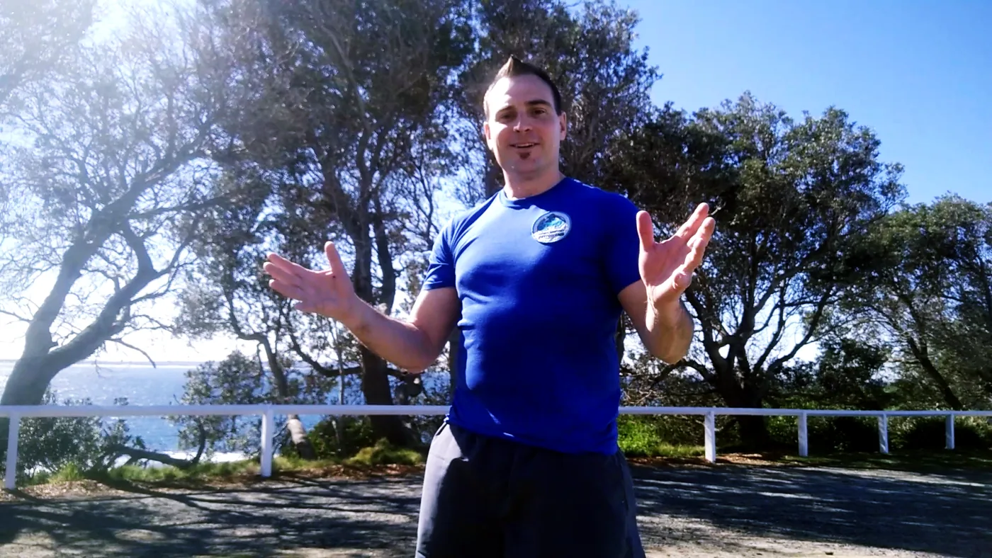 Smiling man in a blue shirt gestures outdoors by a white fence, with trees, ocean, and bright sun in the background.