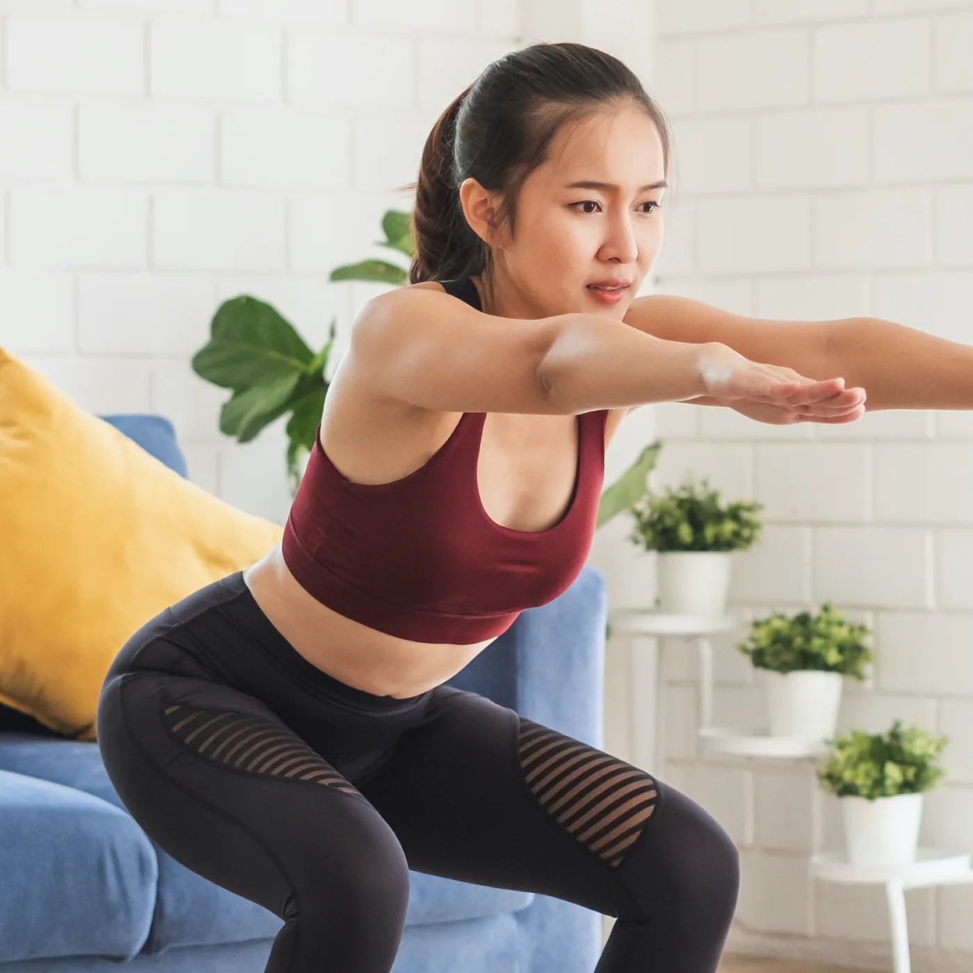 Woman in maroon workout top and black leggings doing a squat in a bright living room with blue sofa, yellow pillow, and plants.