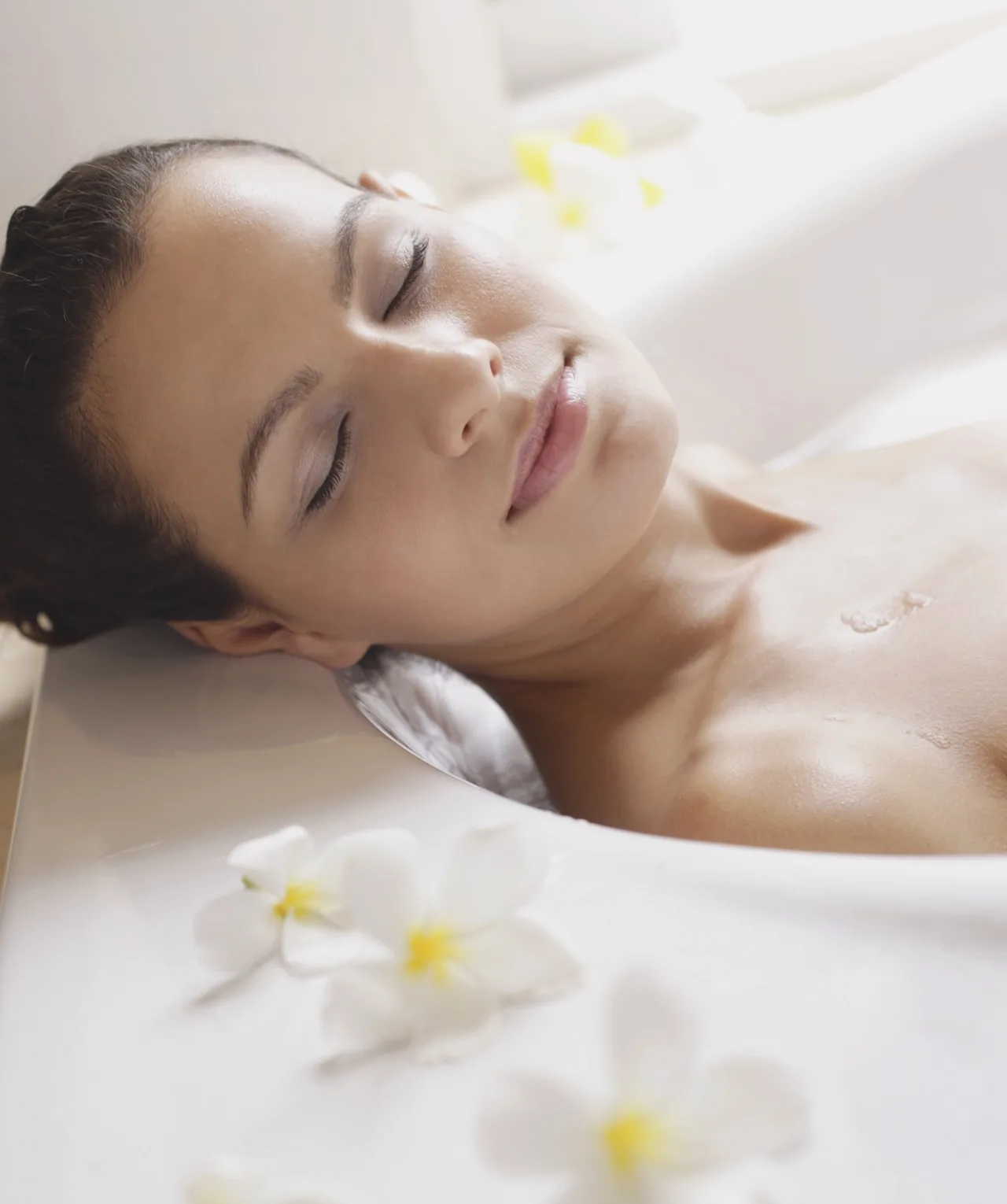 Close-up of a woman with eyes closed relaxing in a white flower-filled bath, serene and spa-like.