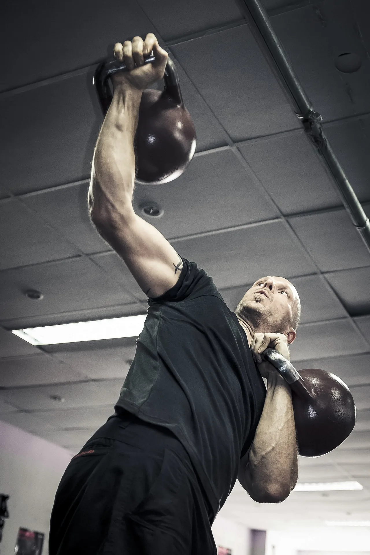 Man in a gym lifts two kettlebells overhead and at his shoulder, focused expression under fluorescent ceiling lights.