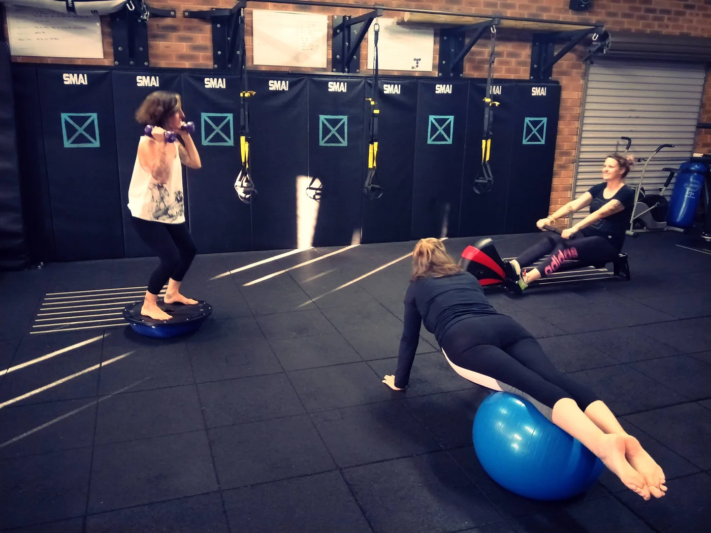 Three women do balance exercises in a gym, one on a wobble board and two on balls, with SMAI mats behind them.
