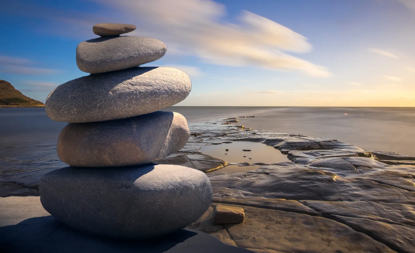 Stacked smooth stones on a rocky shoreline at sunset, with calm sea, soft clouds, and a tranquil golden-blue sky. The stones represent balance.