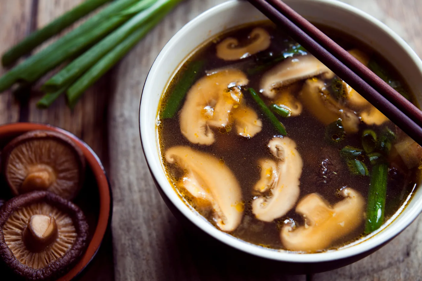 Bowl of mushroom soup with chopsticks, scallions, and sliced mushrooms on a wooden table, close-up.