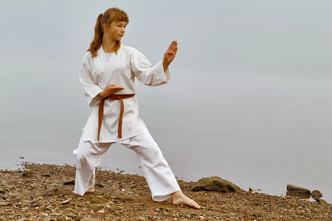 Girl in a white karate gi and brown belt holds a martial arts pose on a rocky lakeshore under a gray sky.