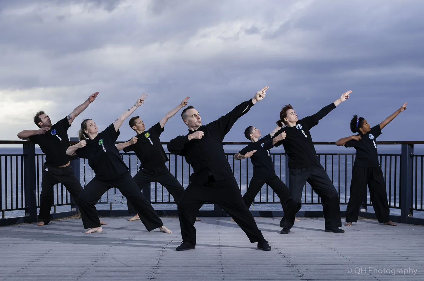 Six martial artists in black uniforms strike synchronized poses on a seaside deck under a cloudy sky.