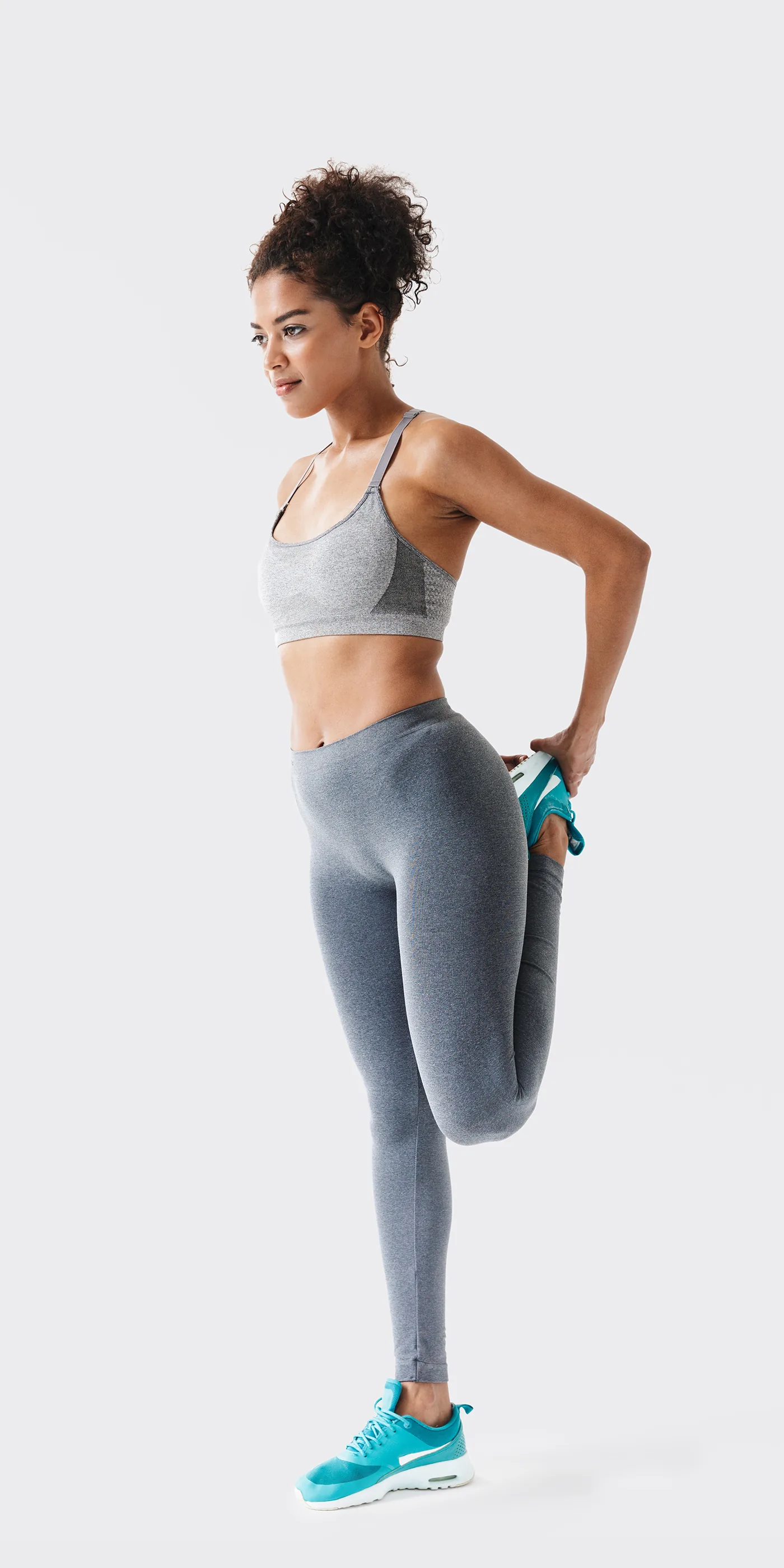 Fit woman in gray athletic wear stretches her quad in turquoise sneakers against a plain white background, focused expression
