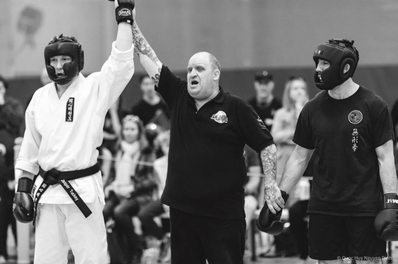 Black-and-white martial arts match: referee raises a fighter’s arm as another headgear-clad opponent stands beside him.