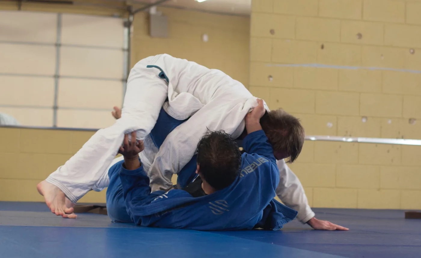 Two ju jitss players grapple on blue mats, one in a white gi lifted over another in a blue gi inside a gym.