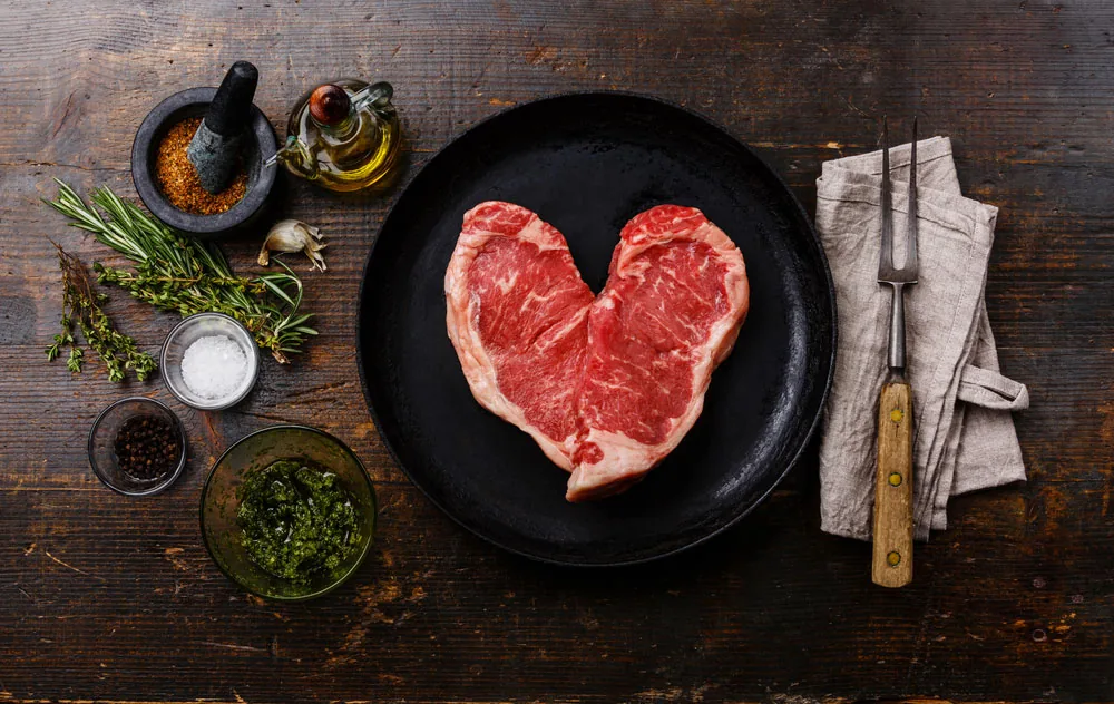 Heart-shaped raw steak on a black plate, surrounded by herbs, salt, oil, and a fork on a napkin over rustic wood.